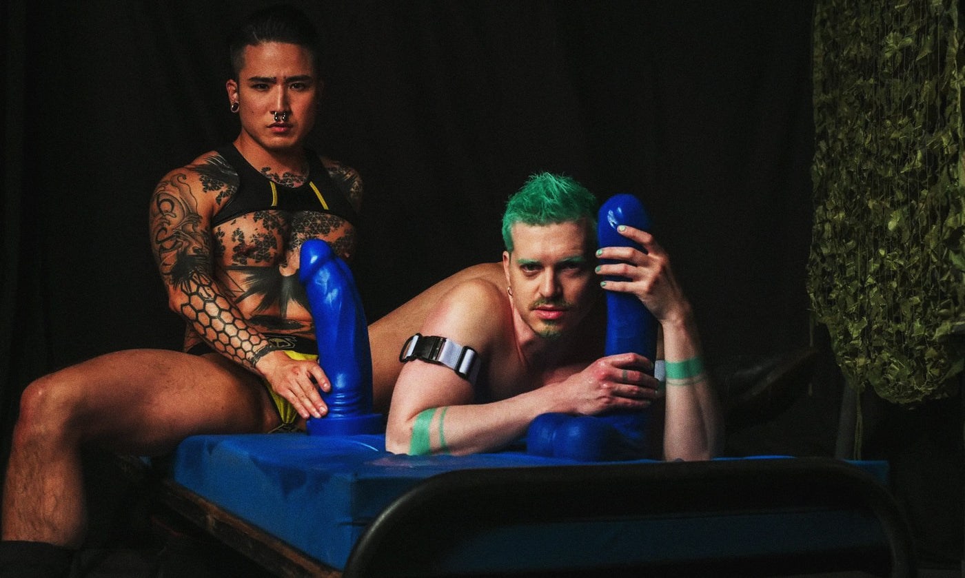 Two people with blue dildos resembling toys or props against a dark background